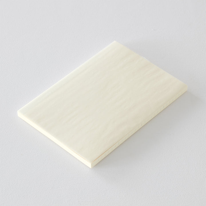 A5 MD Notebook Diary 2026 with a minimalist cream-toned glassine paper cover, thread-stitched binding, and a white bookmark string. The open spread displays a block-style monthly calendar on one side and lined memo pages on the other, printed on smooth MD PAPER designed for a premium writing experience.